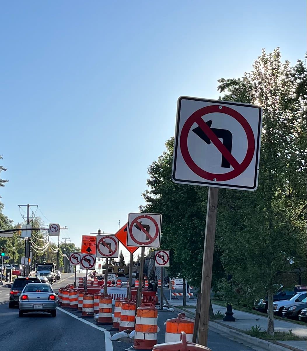 A street scene showing a construction zone with multiple 'No Left Turn' signs and orange traffic barrels lining the road. A silver car is driving away from the camera on the left side of the street.