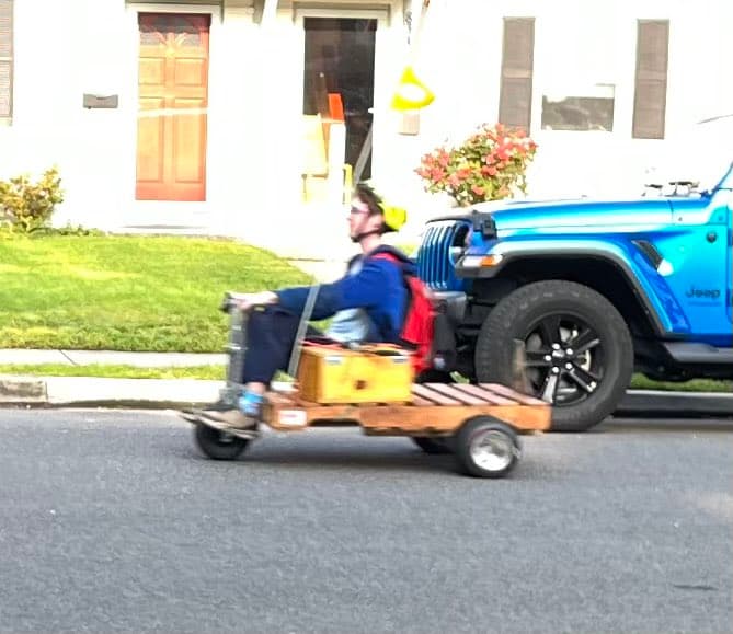 A person wearing a blue shirt and a red backpack is riding a motorized pallet down a residential street, passing a parked blue Jeep. The background features a white house with a red door and a green lawn.