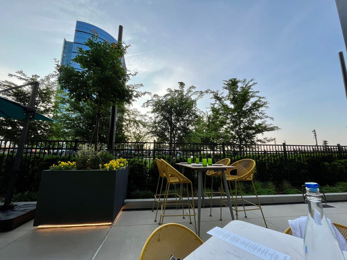 A photo of an outdoor rooftop patio featuring a high-top table with yellow chairs, a planter with yellow flowers, and a view of a modern glass skyscraper in the background.
