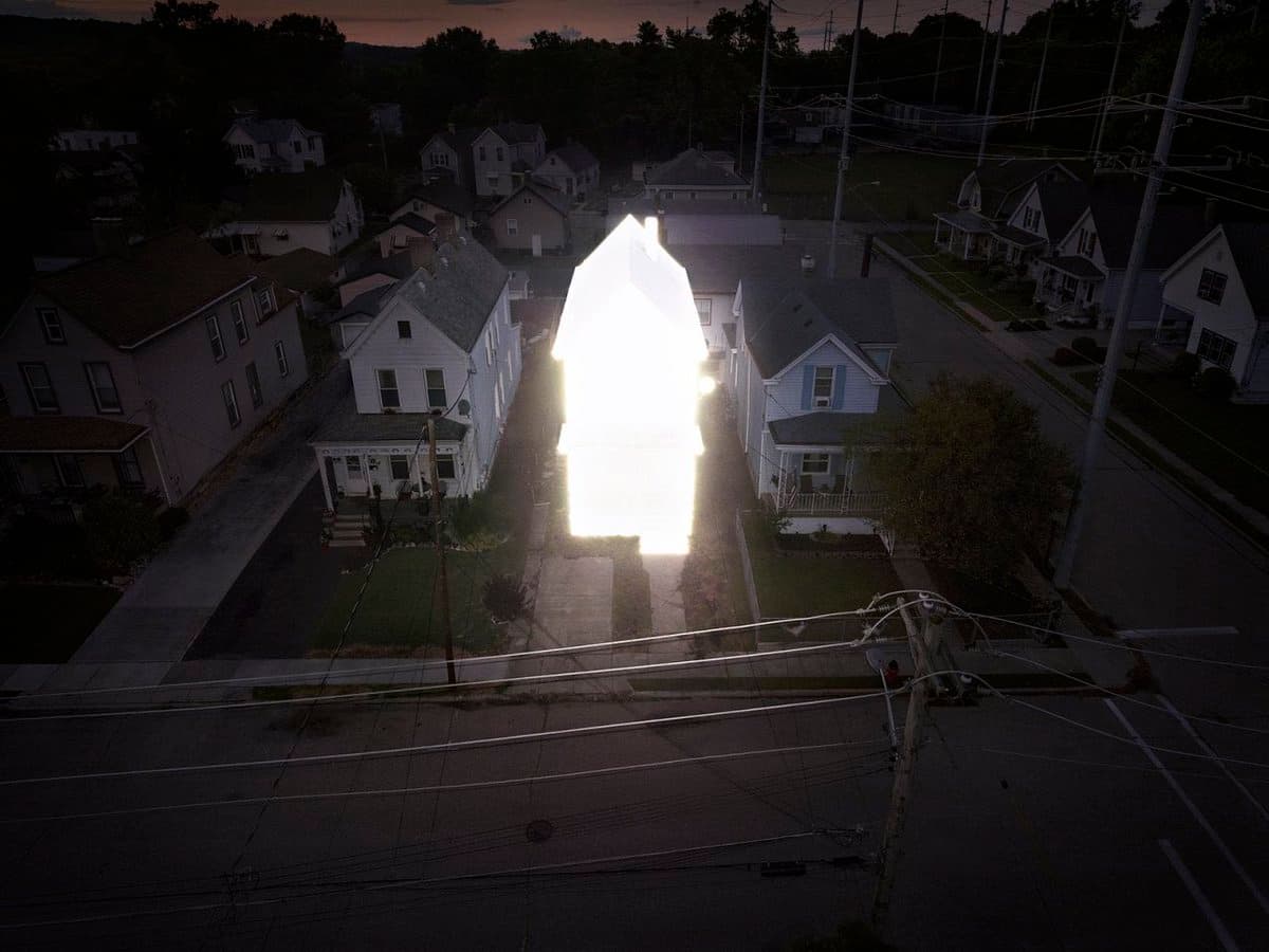 An aerial photograph of a residential neighborhood at dusk, featuring a central house that is glowing with intense, blinding white light, contrasting with the dark surroundings.