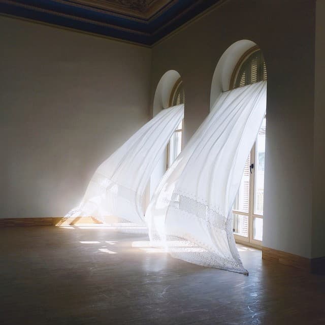 A photograph of a minimalist interior featuring two large, white, lace-trimmed fabric installations billowing out from arched windows onto a wooden floor.