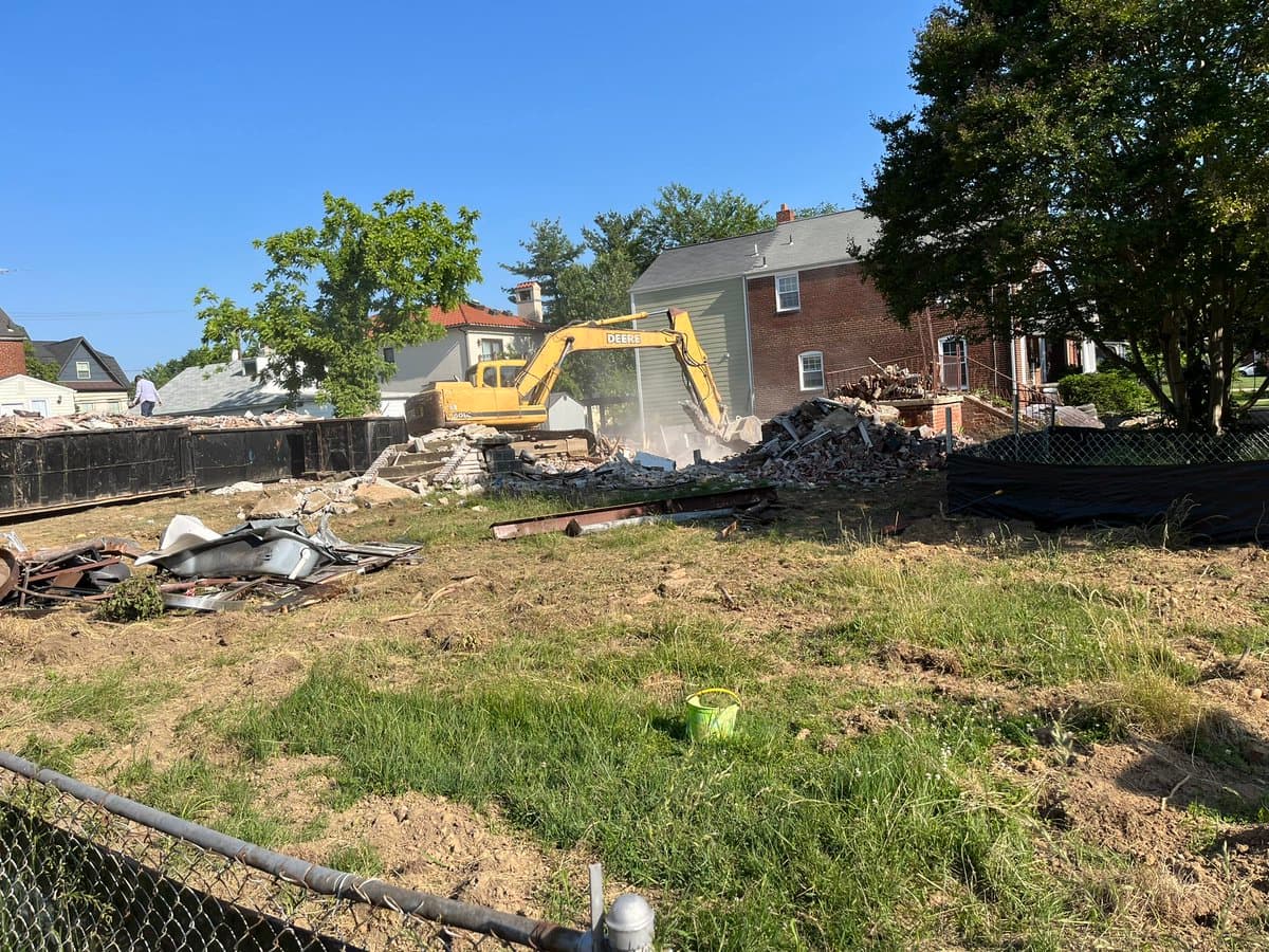 A photograph of a residential demolition site on a sunny day, showing a yellow Deere excavator working on a pile of rubble in front of a brick house. The foreground consists of a grassy, dirt-covered lot with a chain-link fence and a green bucket, while large black dumpsters are visible on the left.