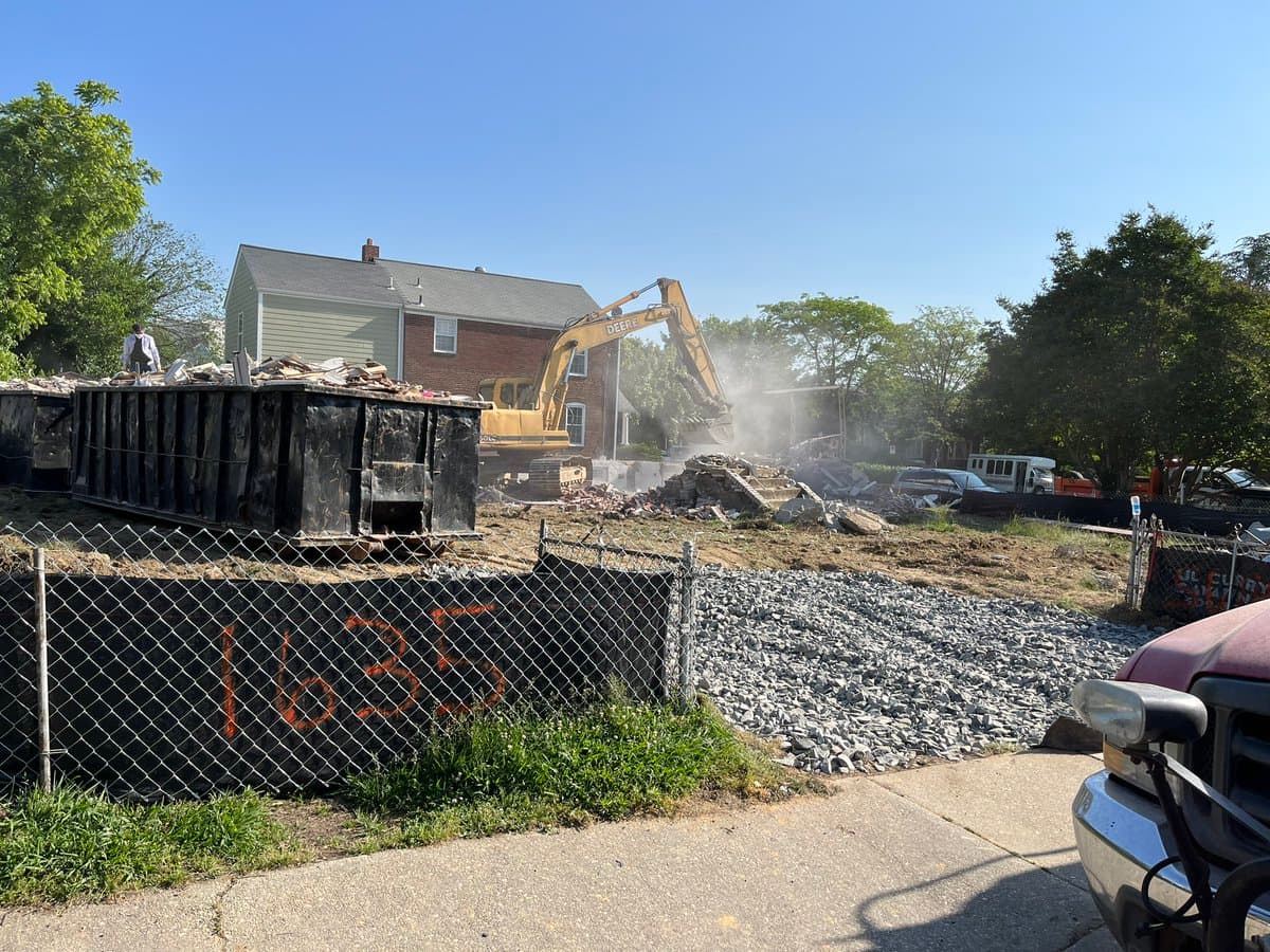 A photograph of a residential demolition site featuring a yellow excavator actively tearing down a brick house, with a large black dumpster in the foreground and a chain-link fence marked with the number 1635.
