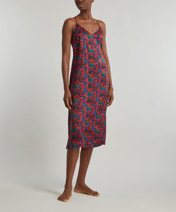 A woman stands barefoot against a plain white background, wearing a sleeveless, knee-length slip dress with a colorful floral pattern in shades of red, blue, and green. The dress features thin spaghetti straps and a V-neckline.