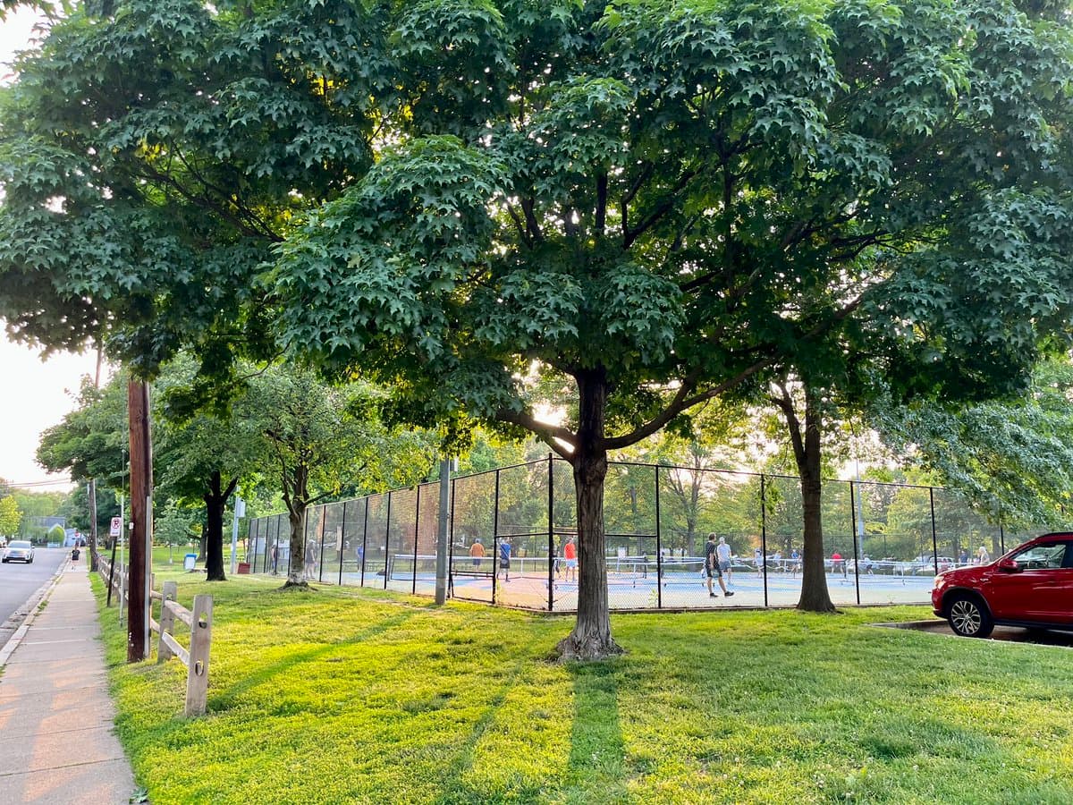 A photograph of an outdoor tennis court complex surrounded by a chain-link fence, with several people playing on the blue and green courts. The scene is framed by large, leafy trees and a grassy area, with a sidewalk and a red car visible in the foreground.