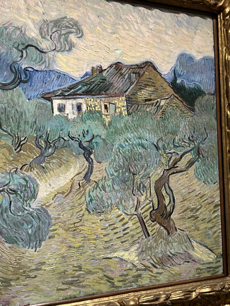A photograph of a framed oil painting depicting a rural landscape with a house, olive trees, and mountains, rendered in a thick, expressive impasto style.
