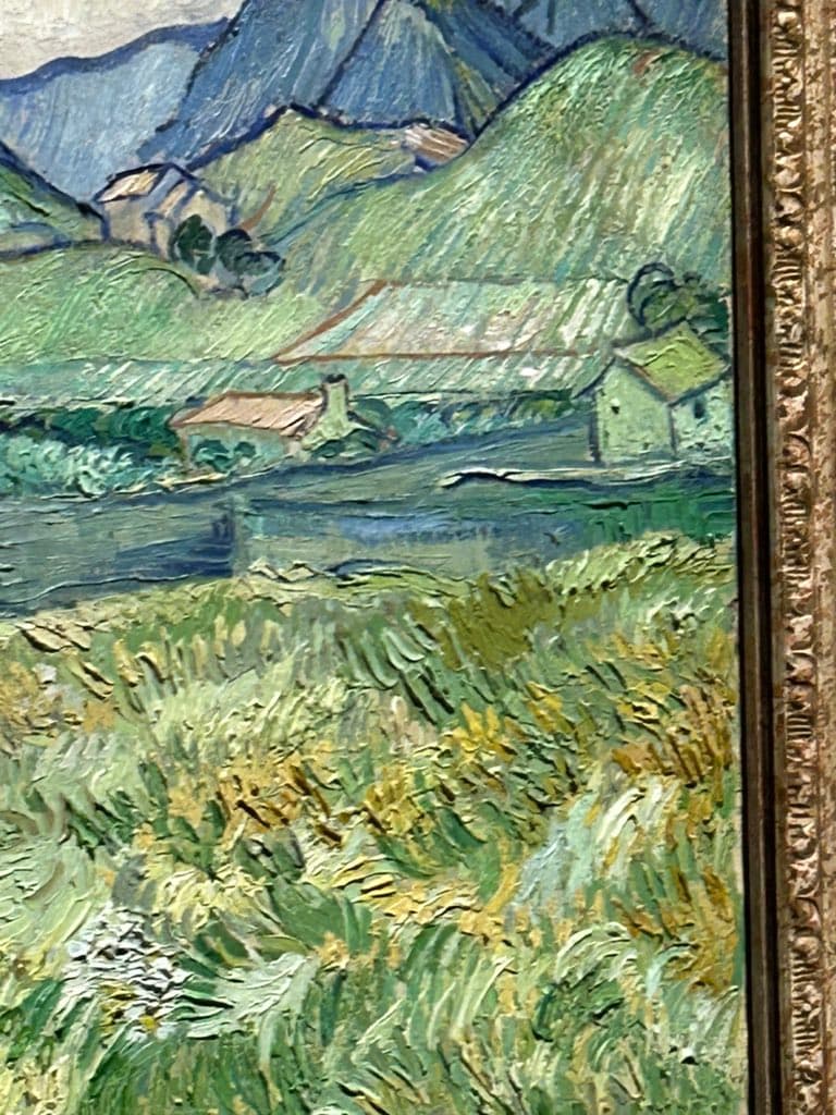 A close-up photograph of a framed oil painting depicting a landscape with rolling green hills, small houses, and a body of water, rendered in a thick, textured impasto style.