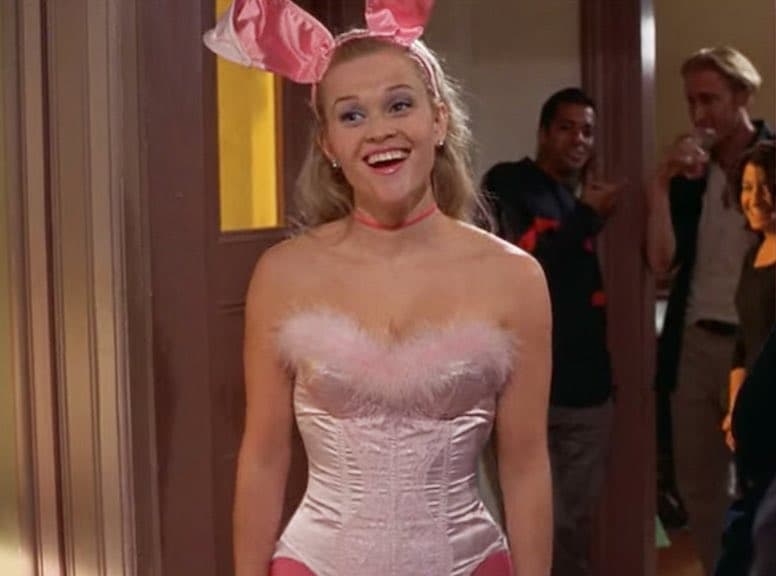 A still from a movie or TV show featuring a smiling woman in a pink costume with bunny ears, standing in a doorway with other people in the background.