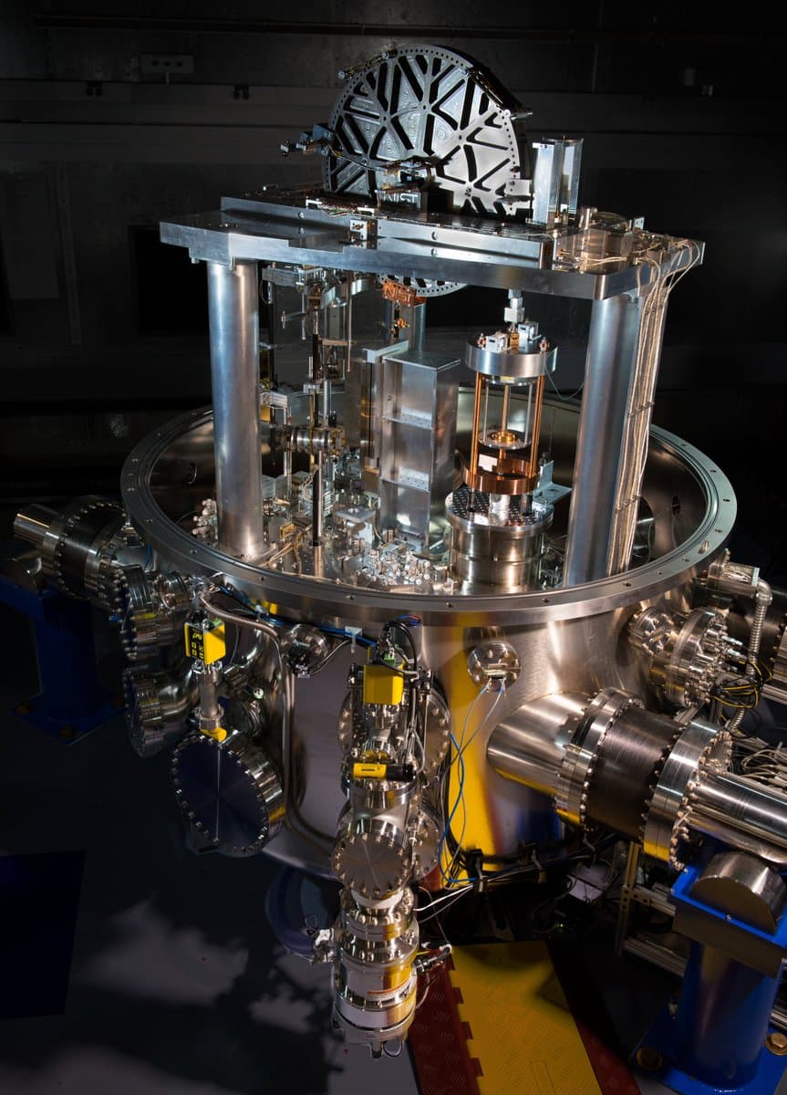 A large, complex piece of scientific equipment, likely a particle accelerator or vacuum chamber, featuring polished metal pipes, cylindrical components, and a circular top structure with a spoked design. The device is mounted on blue support stands in a dimly lit laboratory setting.