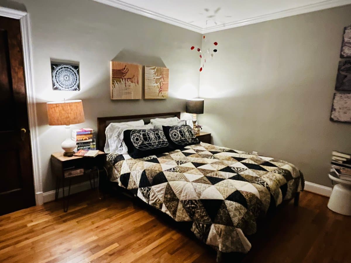 A photograph of a cozy bedroom featuring a bed with a black, white, and brown geometric patchwork quilt, flanked by two nightstands with lamps and books. The room has light gray walls, hardwood floors, and wall art including a dreamcatcher and abstract pieces.