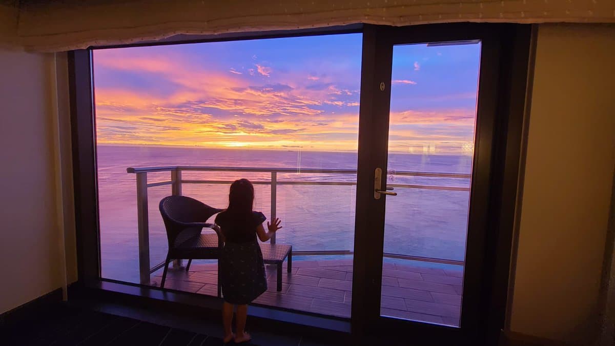 A young child stands on a balcony looking out at a vibrant sunset over the ocean. The view is framed by a glass sliding door, with a wicker chair visible on the deck.