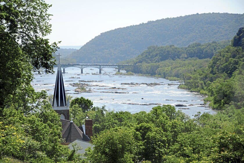 A scenic landscape photograph showing a wide river flowing through a lush, green valley with a bridge in the distance and a church steeple in the foreground.