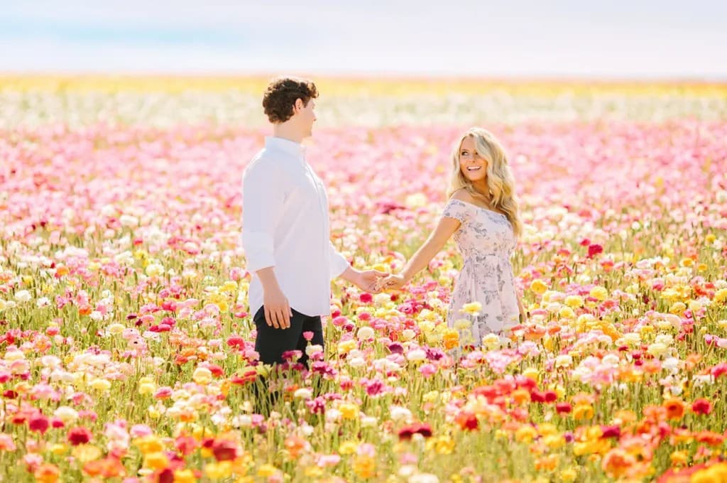 A couple holding hands in a vast field of colorful pink, yellow, and red flowers on a sunny day. The man wears a white button-down shirt and dark pants, while the woman wears a light-colored floral dress.