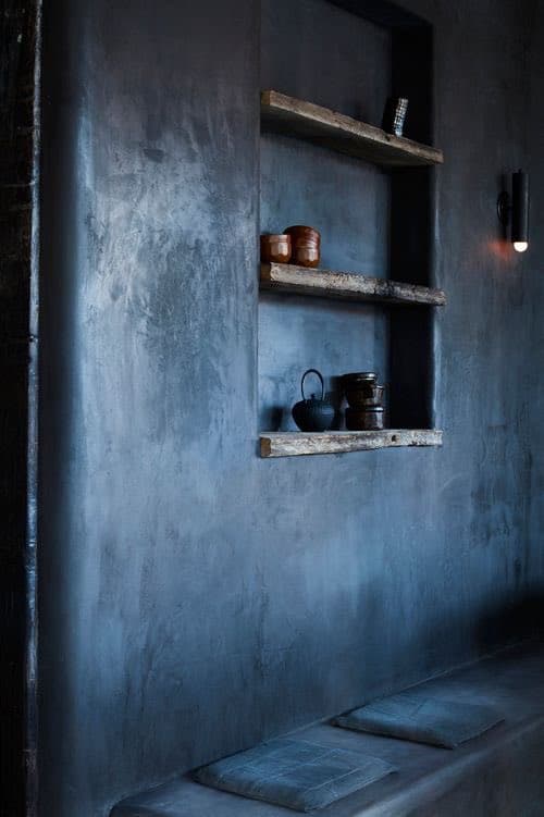 A dimly lit interior space featuring a dark, textured wall with three rustic wooden shelves holding ceramic pots and a small teapot. Below the shelves, a low bench is fitted with two square cushions, and a wall sconce provides soft lighting on the right.