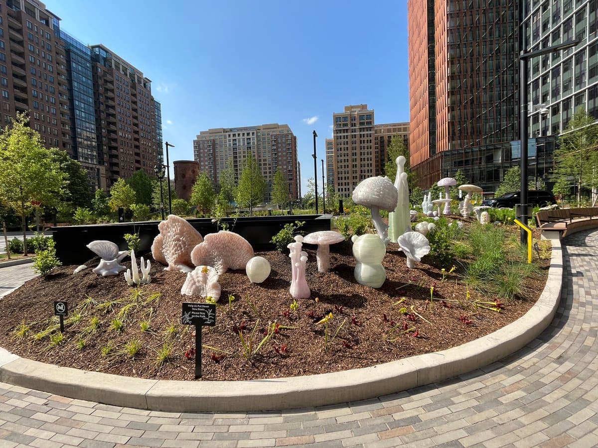 A photo of a landscaped garden bed in an urban plaza, featuring white and pink ceramic sculptures shaped like mushrooms and coral, surrounded by mulch and small plants. Tall apartment buildings and a clear blue sky are visible in the background.