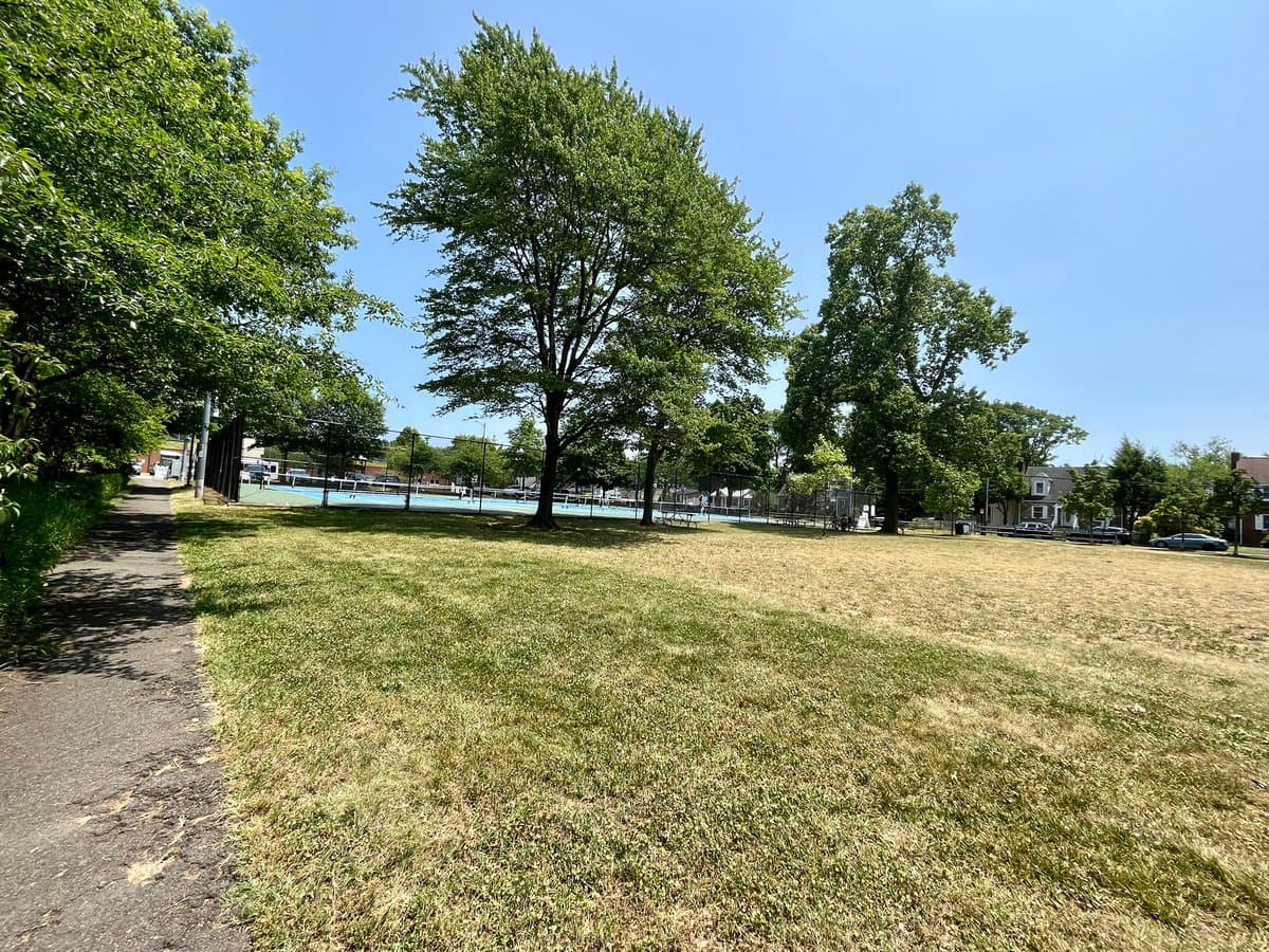A wide outdoor photograph of a park on a sunny day, featuring a grassy field in the foreground and a paved path on the left. In the background, there are several tennis or pickleball courts surrounded by a chain-link fence, with large green trees and houses visible beyond the courts under a clear blue sky.