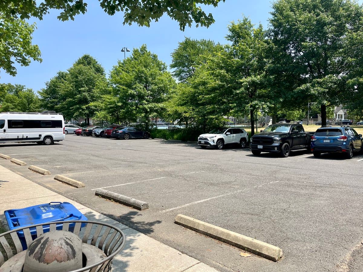 A daytime outdoor photograph of a paved parking lot with several cars and a white van parked under a line of green trees, with a blue recycling bin and a metal trash can in the foreground.
