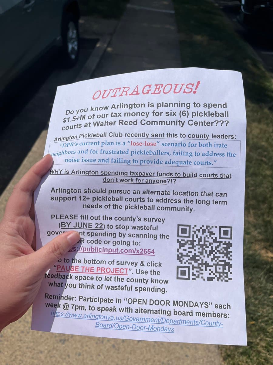 A close-up photograph of a person's hand holding a printed flyer titled 'OUTRAGEOUS!' in red text. The flyer is held outdoors on a sidewalk, with a parked car and grass visible in the background. The document contains text opposing a local government plan to build pickleball courts and includes a QR code.