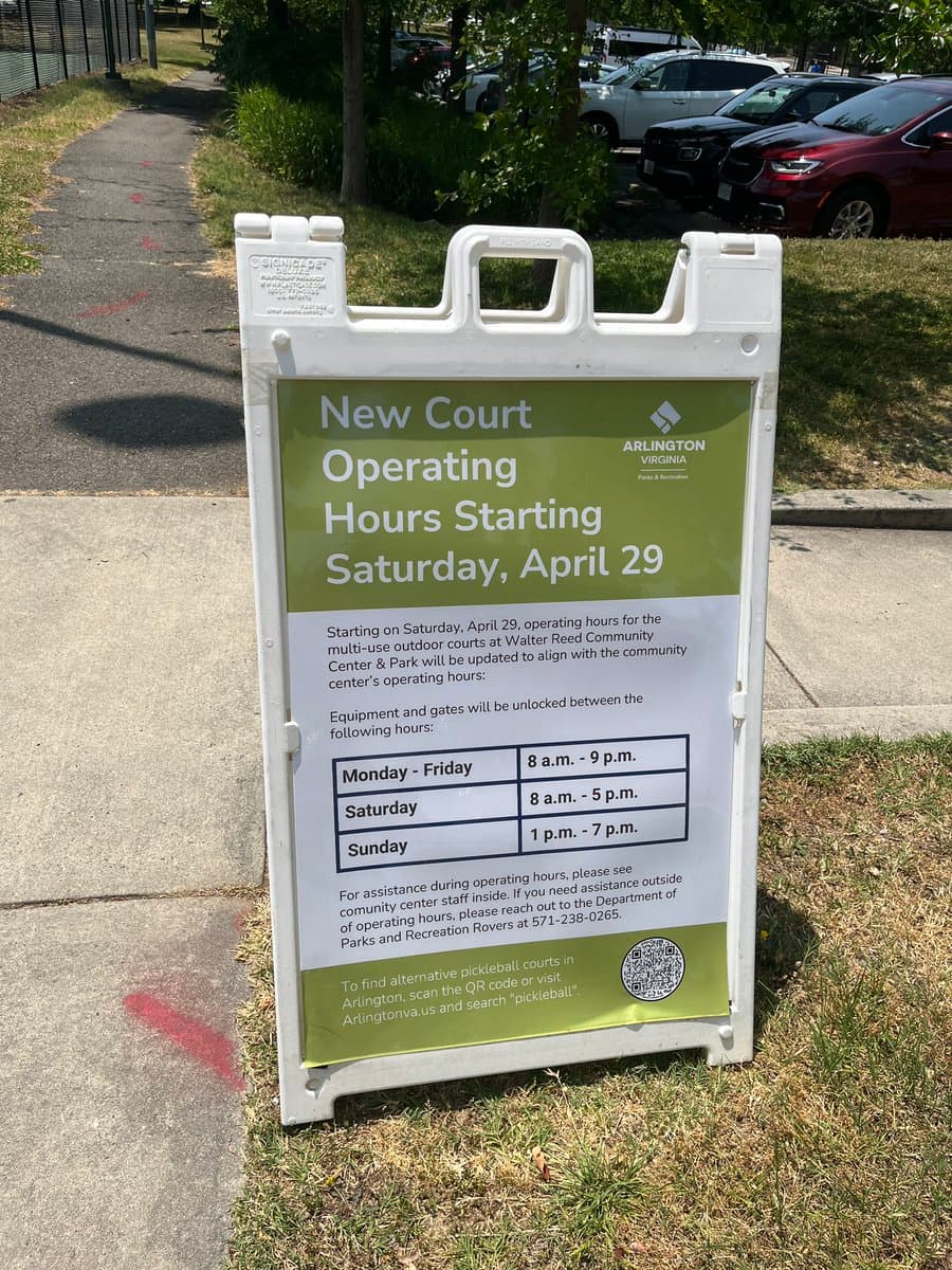A white A-frame sign on a grassy area near a sidewalk announces new operating hours for outdoor courts at the Walter Reed Community Center & Park in Arlington, Virginia. The sign includes a schedule table and a QR code for finding alternative pickleball courts.