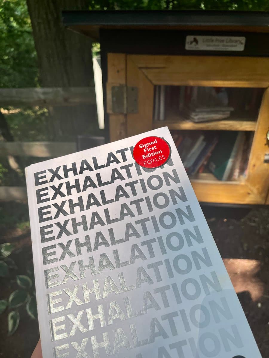 A close-up photograph of a person holding a copy of the book "Exhalation" in front of a wooden Little Free Library. The book cover features the title repeated in a fading, metallic font and has a red sticker indicating it is a signed first edition from Foyles.
