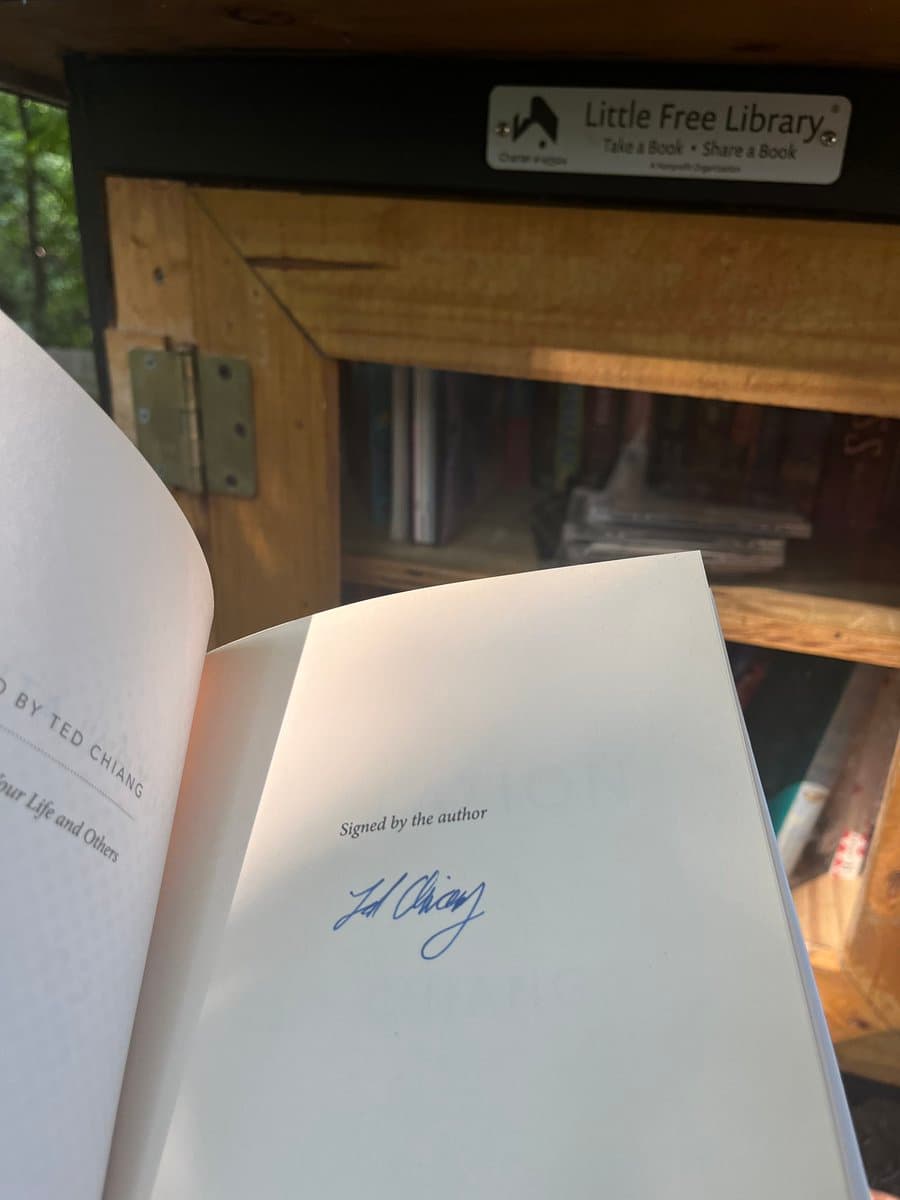 A close-up photograph of an open book held in front of a wooden Little Free Library box. The book's title page shows the author's name, Ted Chiang, and a handwritten signature in blue ink below the printed text "Signed by the author".