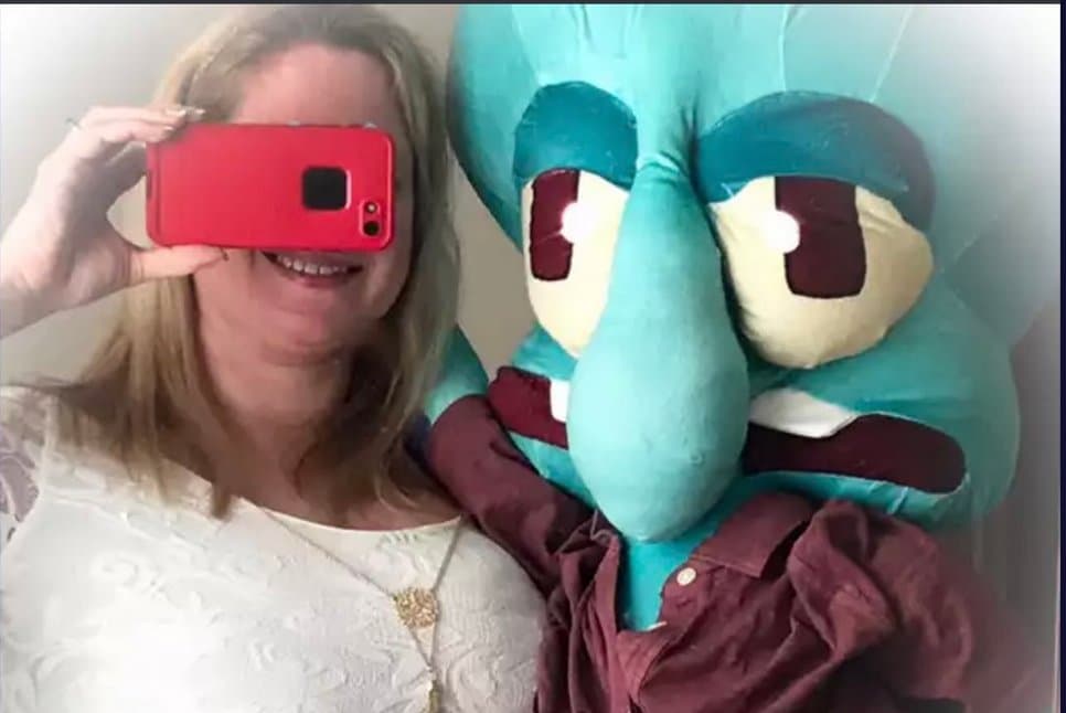 A woman holding a red smartphone takes a selfie next to a large Squidward plush toy wearing a maroon shirt.