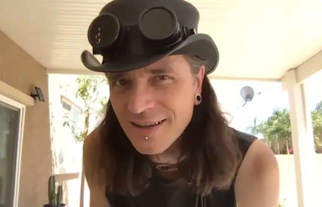 A person with long dark hair is wearing a black top hat with goggles attached to the front, standing on a covered porch with a house and trees visible in the background. The individual is smiling and looking toward the camera.