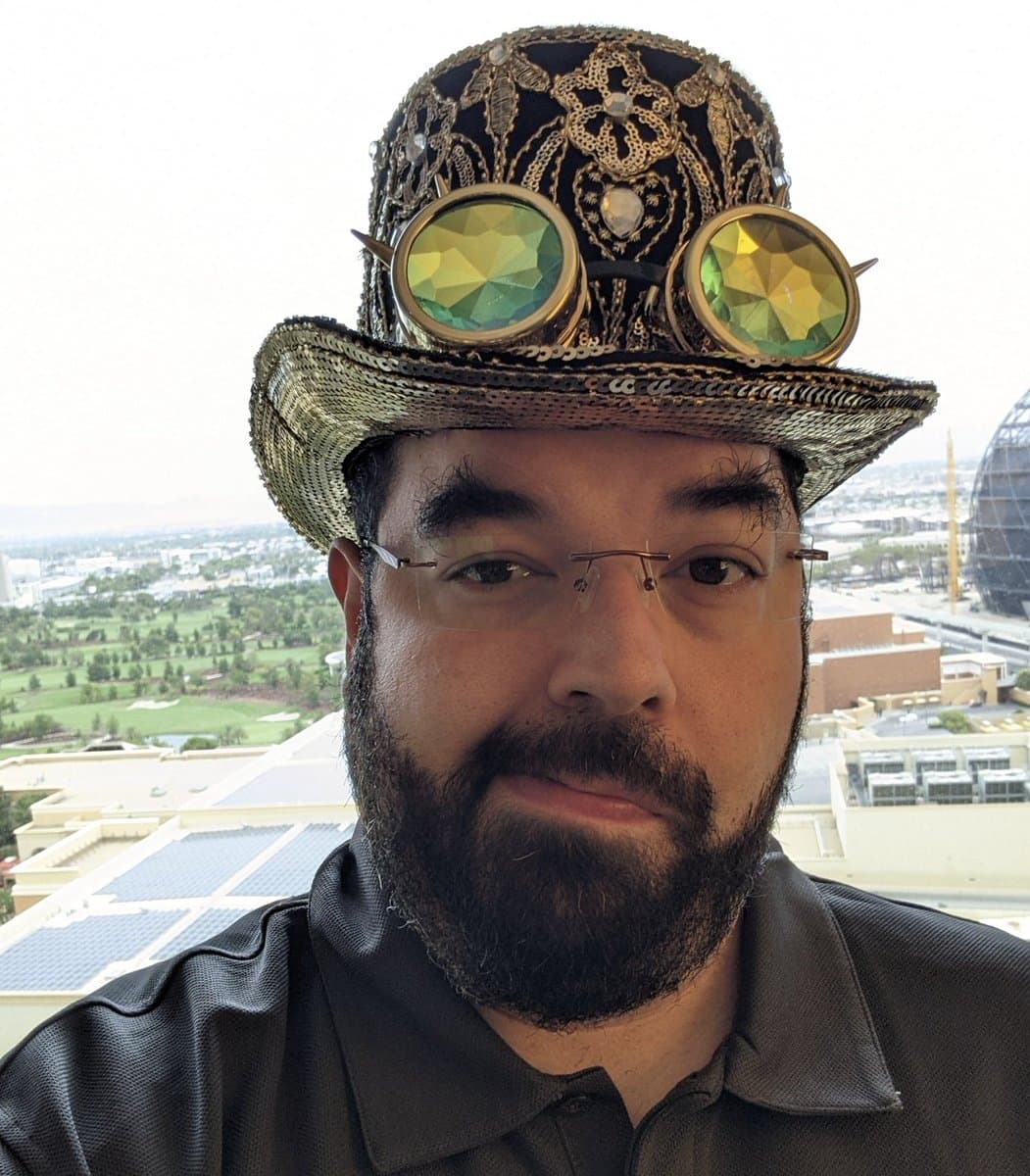 A man with a beard and rimless glasses wears an ornate, steampunk-style top hat decorated with gold filigree and two large, faceted green lenses. He is photographed outdoors against a backdrop of a cityscape and a golf course.