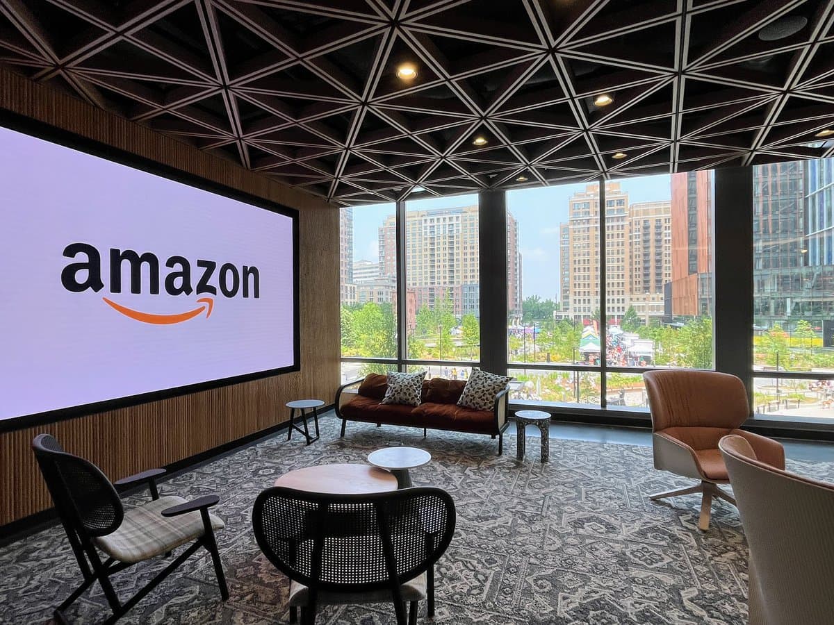 A modern office lounge or meeting space with a large wall-mounted screen displaying the Amazon logo, contemporary furniture, and floor-to-ceiling windows overlooking a city skyline. The room features a geometric ceiling design and a patterned carpet.