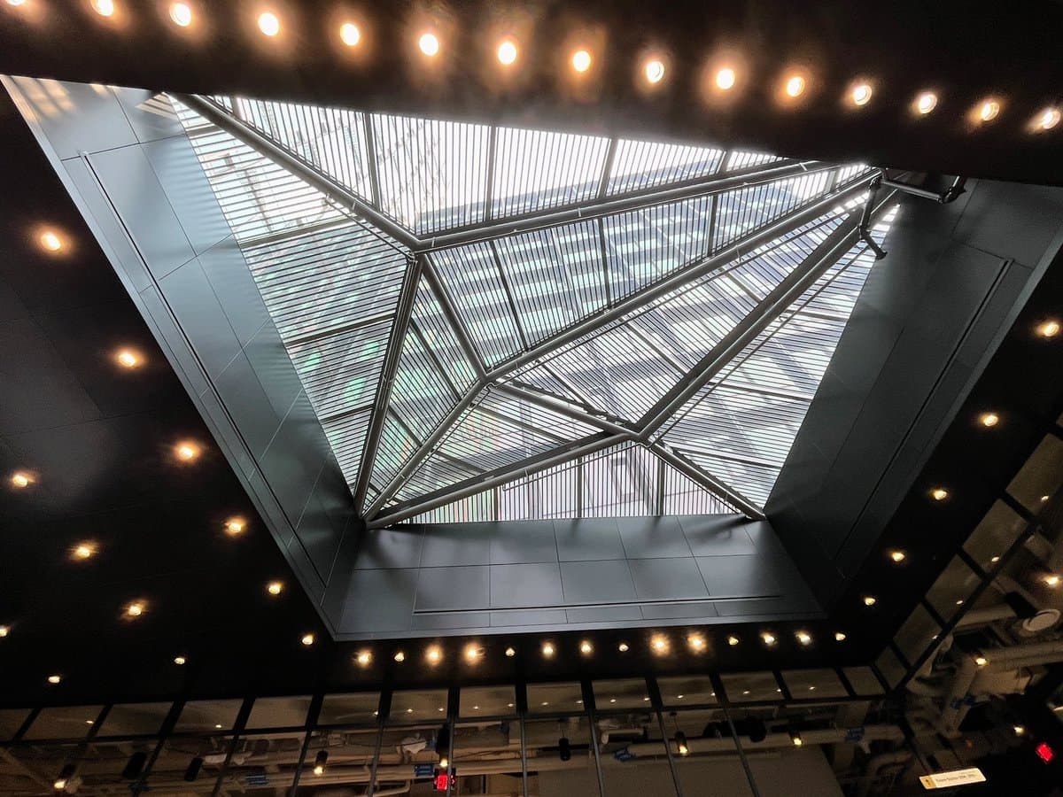A low-angle photograph looking up into a large, modern architectural atrium featuring a geometric glass and steel skylight. The ceiling is lined with numerous recessed lights, and the structure is framed by dark, paneled walls.