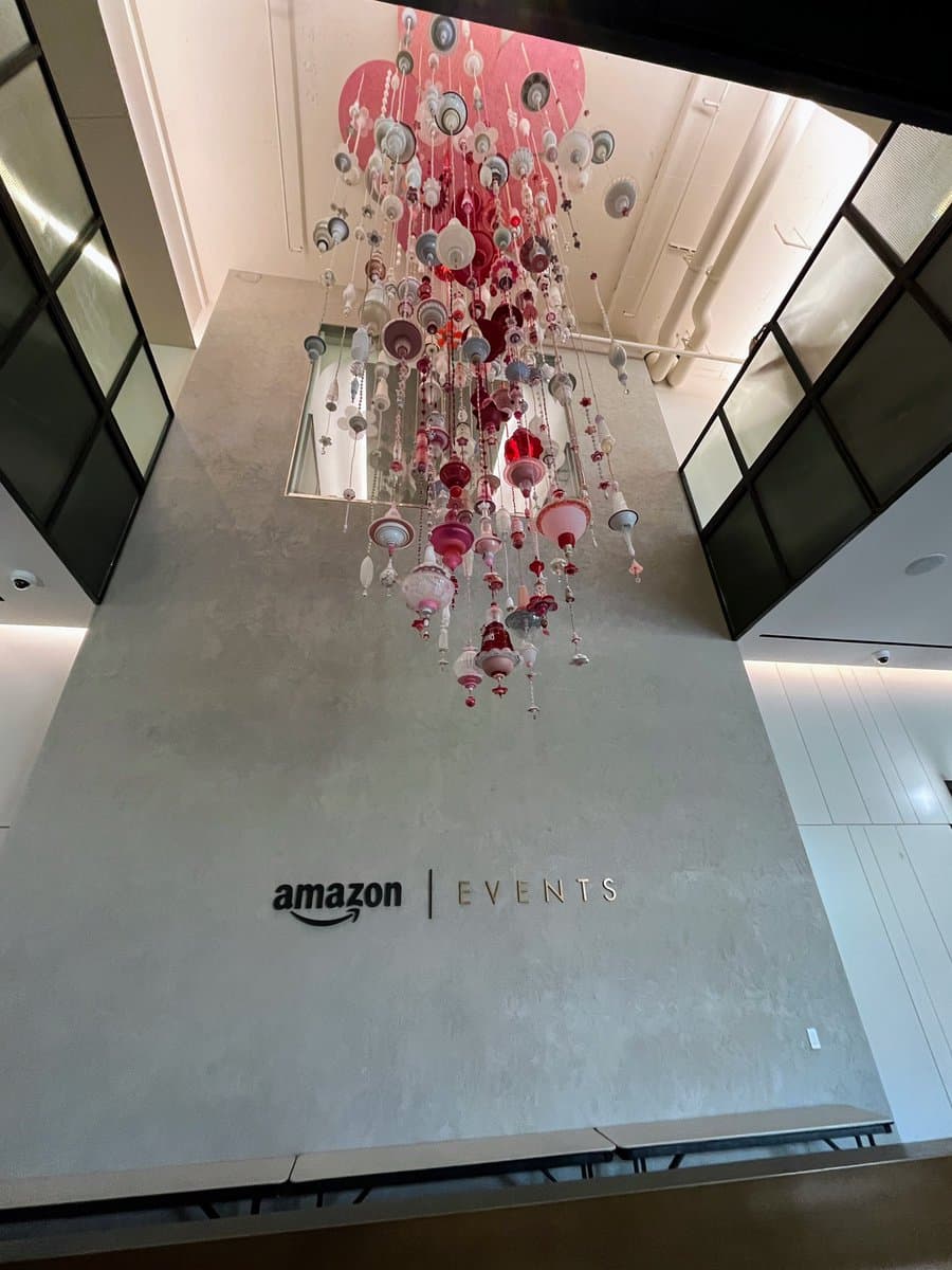 A low-angle photograph of a modern interior space featuring a large, elaborate chandelier made of red and white glass pieces hanging from the ceiling. Below the chandelier, the Amazon logo and the word "EVENTS" are displayed on a concrete wall.