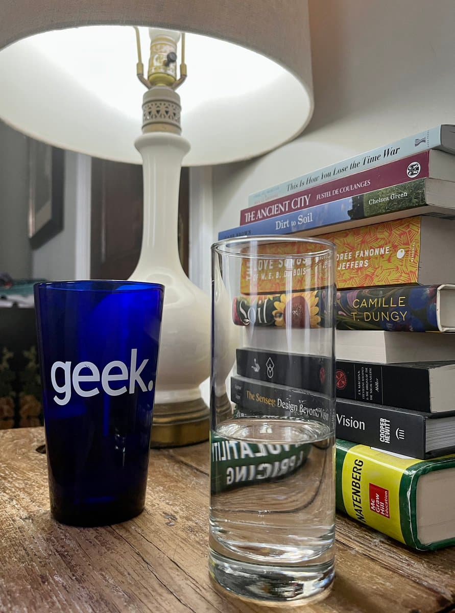 A photograph of a blue glass labeled 'geek.' and a clear glass of water sitting on a wooden table next to a stack of books and a lamp. The books include titles such as 'The Ancient City,' 'Dirt to Soil,' and 'Camille T. Dungy.'