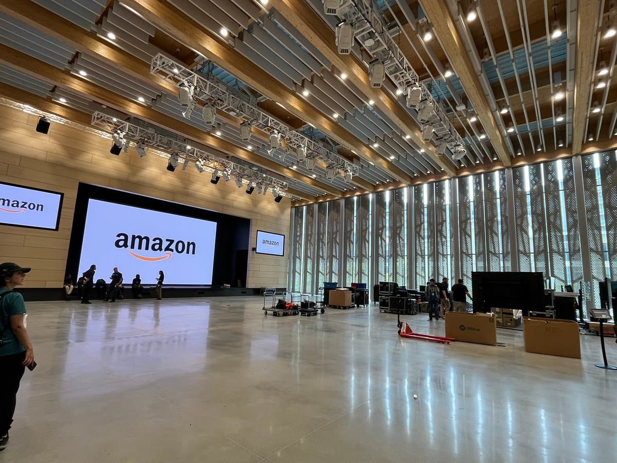 A wide-angle photograph of a large, empty conference hall with a polished concrete floor and a high ceiling featuring exposed wooden beams and lighting rigs. Large screens on the far wall display the Amazon logo, while several people and equipment carts are scattered throughout the space.