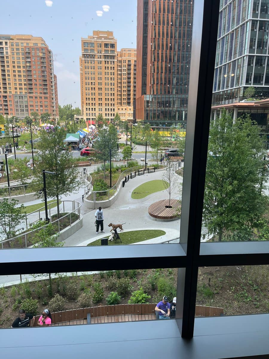 A view from a high-rise window looking down onto a modern urban park with winding pathways, green spaces, and trees. In the background, a busy farmers market with colorful tents is visible, surrounded by tall residential and commercial buildings.