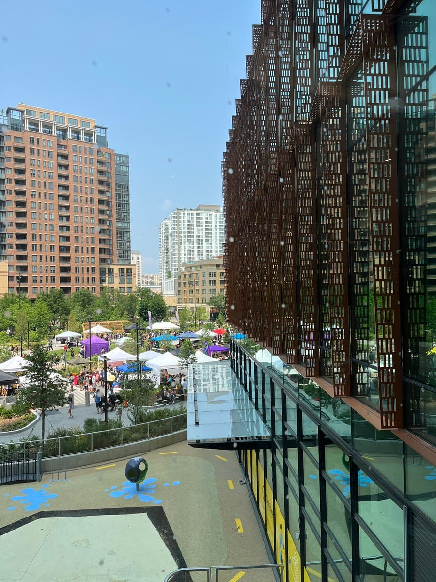 A high-angle outdoor photograph taken from a balcony overlooking a city park with a busy farmers market, featuring colorful vendor tents and people. In the background, several tall residential buildings stand against a clear blue sky, while the foreground shows a playground area with blue painted markings and a modern building with a brown lattice facade on the right.