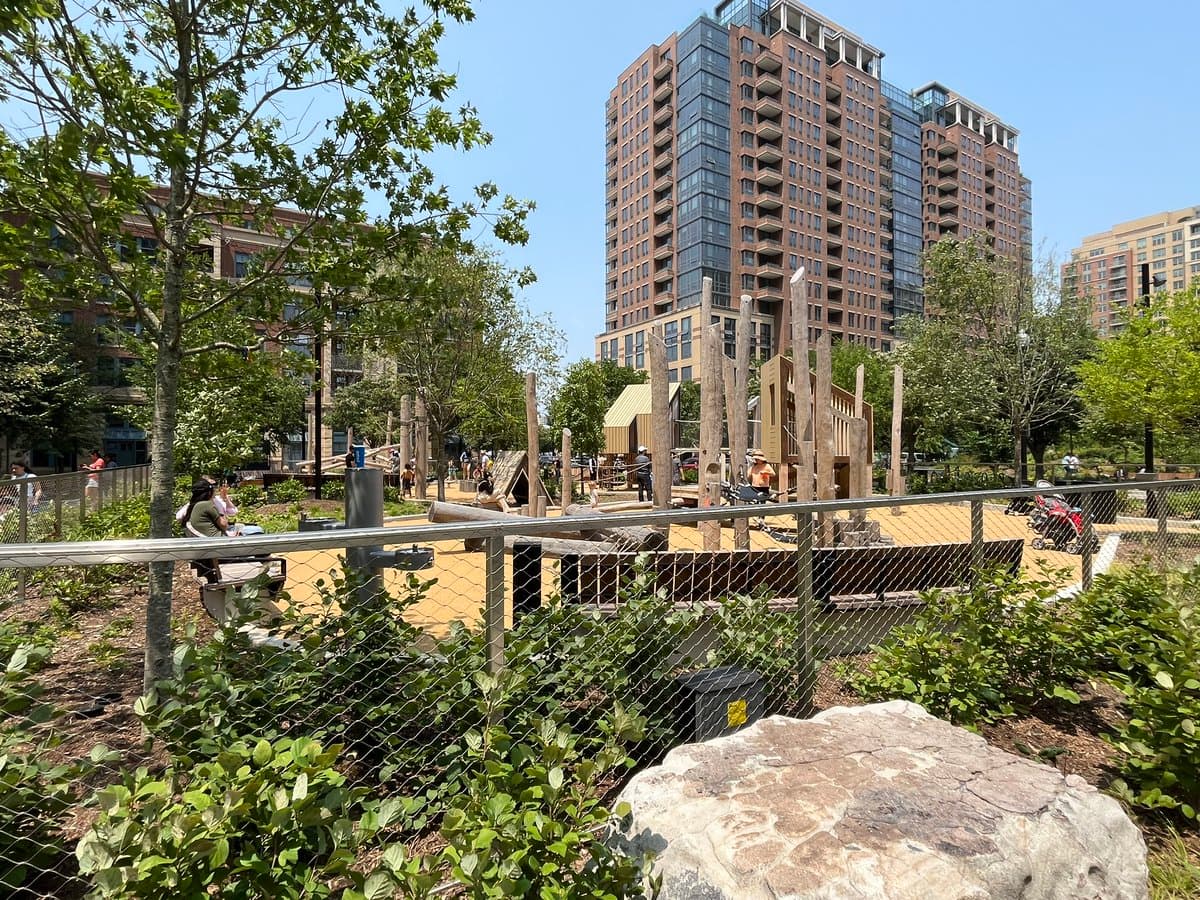 A sunny outdoor view of a city park featuring a wooden children's playground with climbing structures, surrounded by greenery and a chain-link fence, with tall residential buildings in the background.