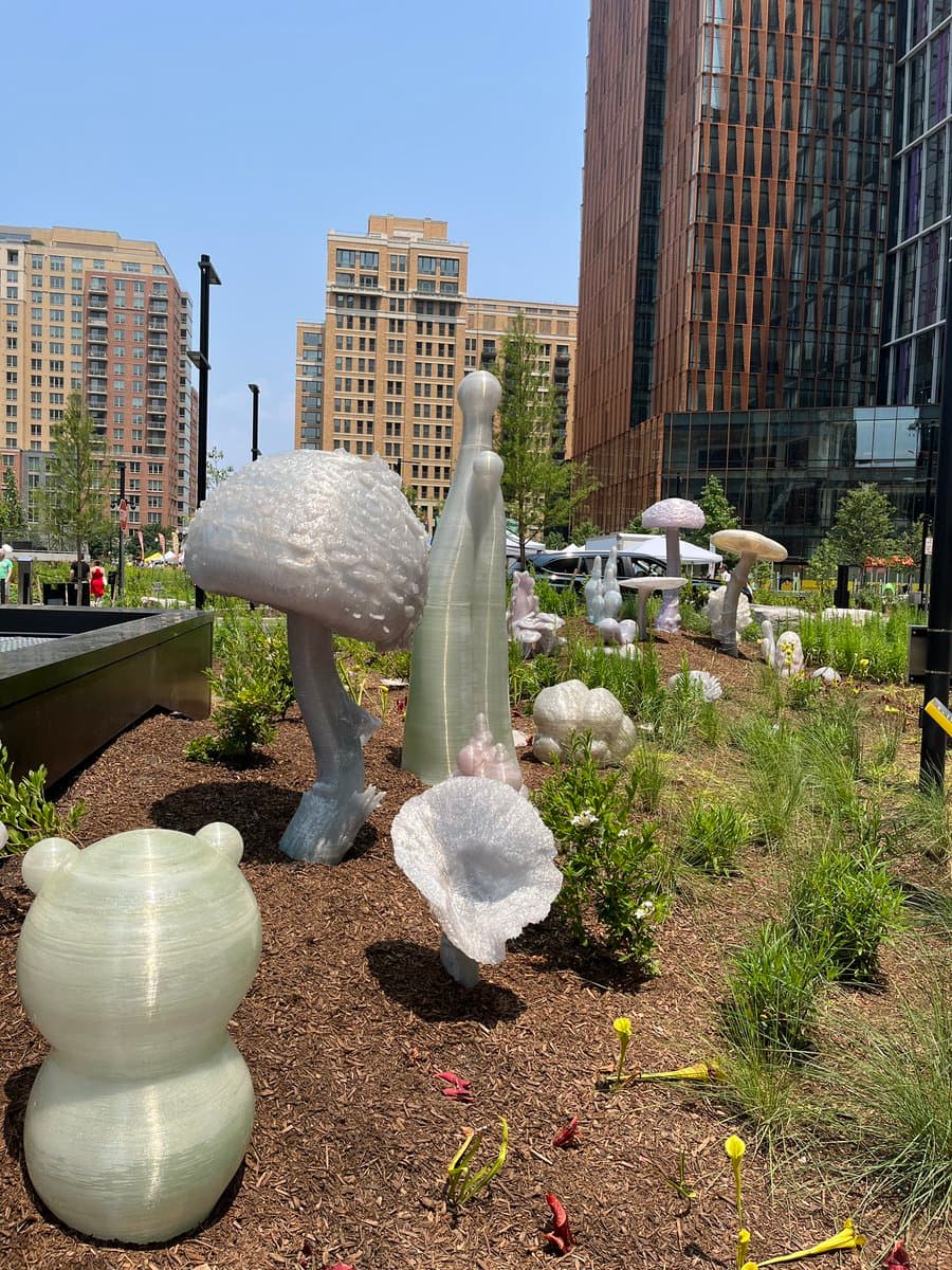 A photograph of a public park garden featuring several large, white, 3D-printed sculptures of mushrooms and a bear-like figure planted in mulch, with tall city buildings in the background.