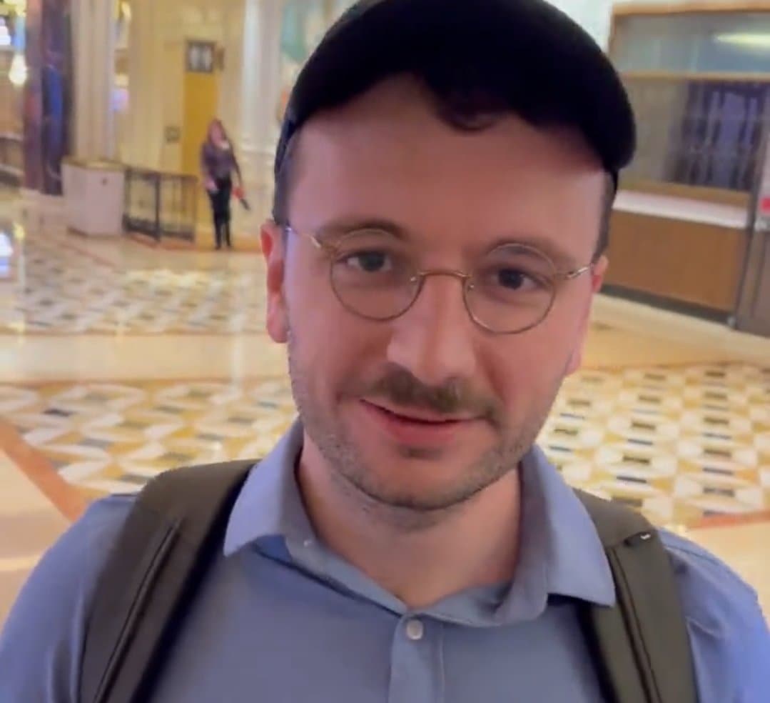 A selfie of a man with short dark hair, a mustache, and round glasses, wearing a light blue collared shirt and a backpack, taken in a brightly lit indoor space with a patterned tile floor. Another person is visible in the background.