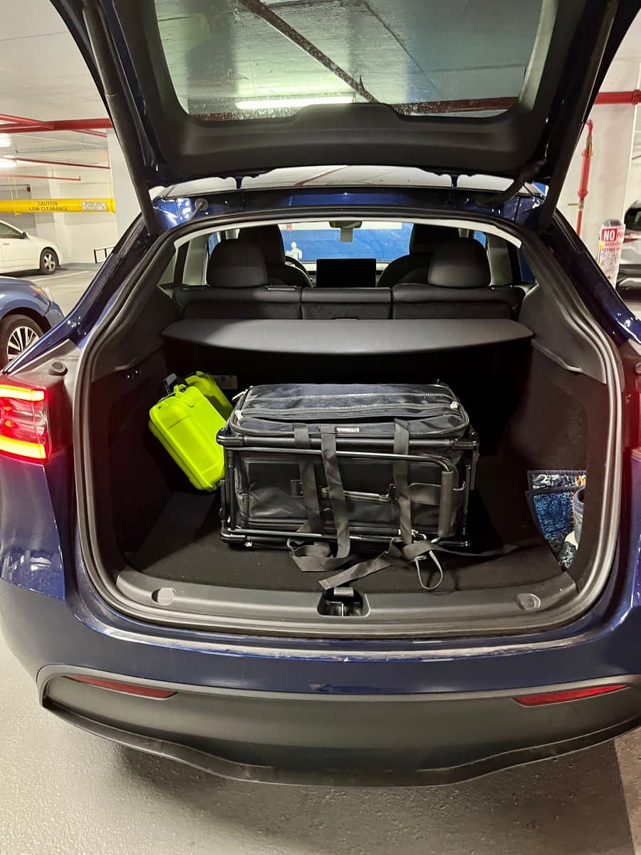 A photo showing the open trunk of a blue car in a parking garage, containing a black folding cart and a green container. The car's rear seats and a laptop are visible through the open hatch.