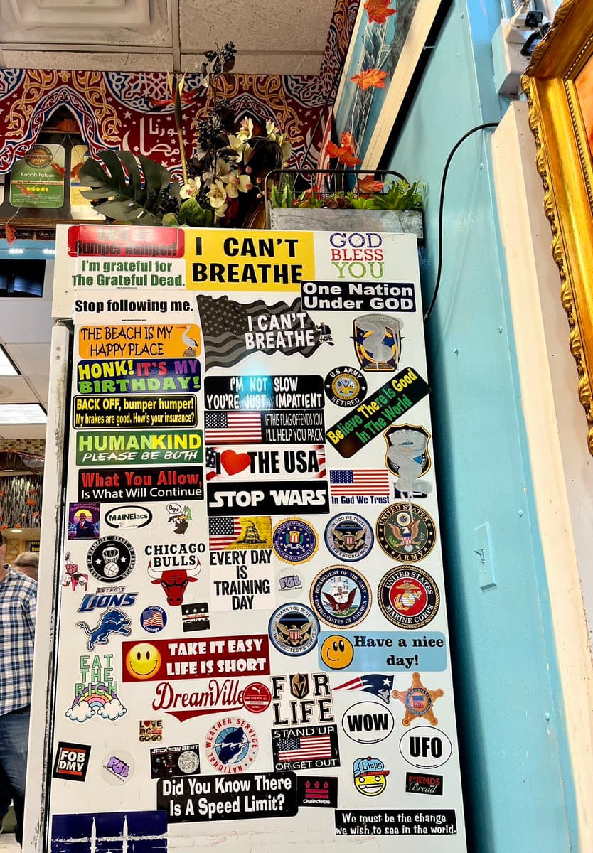 A white surface, likely a refrigerator or door, is covered with a dense collection of bumper stickers and decals. The stickers feature a variety of themes, including patriotic slogans, military branch insignia, sports team logos, and humorous or motivational phrases.