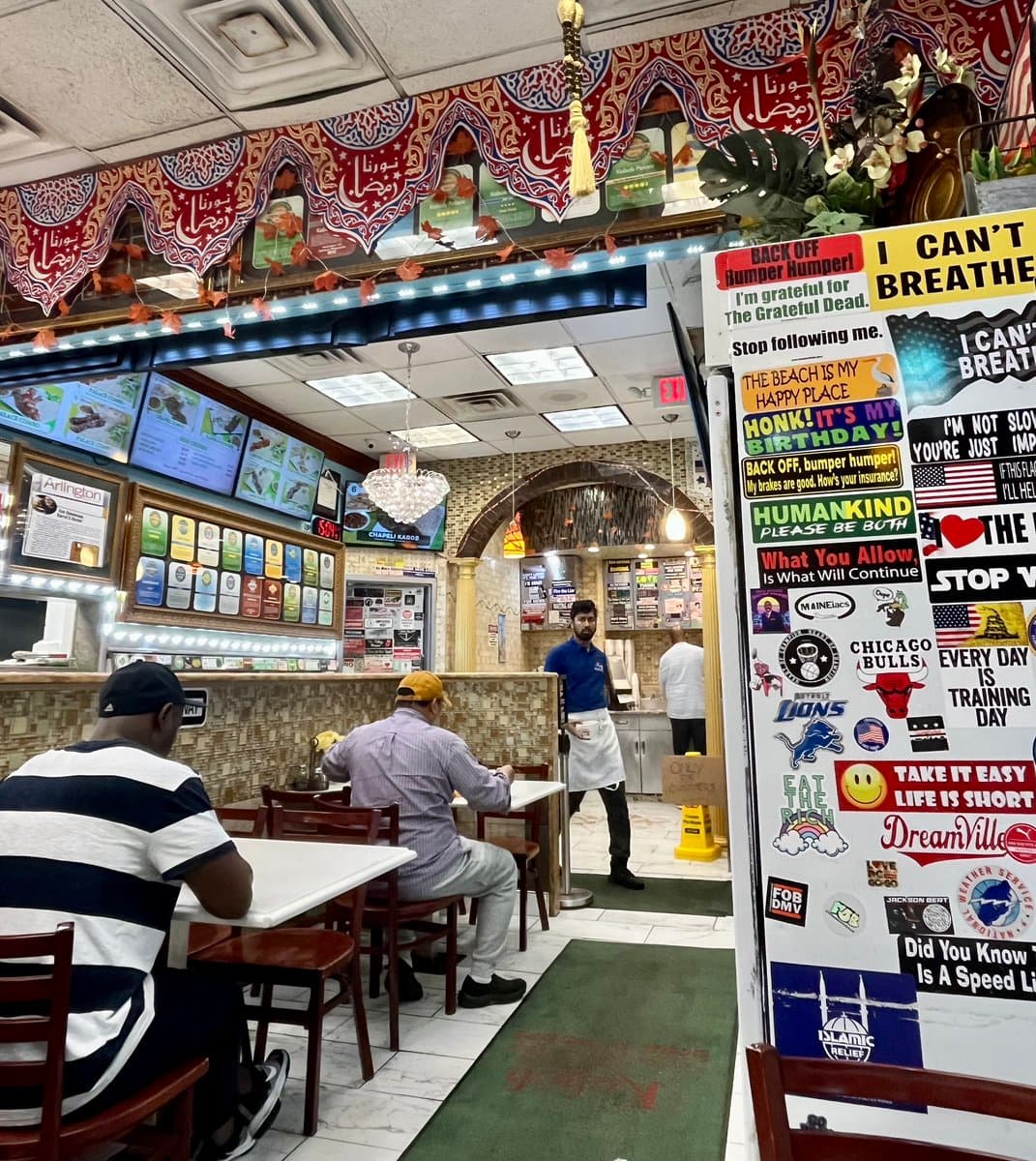 A photograph of the interior of a restaurant featuring a pillar covered in bumper stickers and a decorative red and white valance with Arabic calligraphy. Two customers are seated at a table in the foreground while a staff member stands near the service counter in the background.