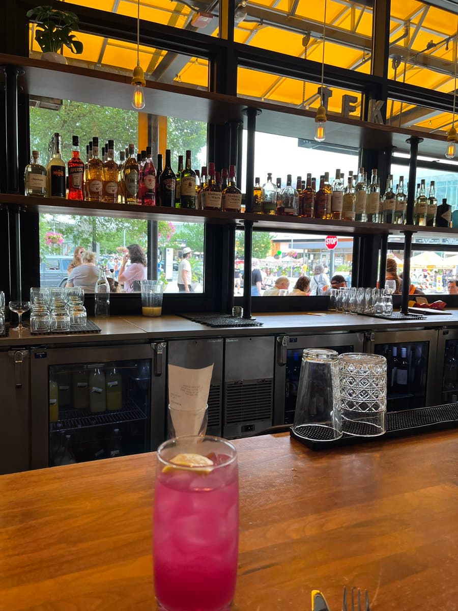 A view from a wooden bar counter looking out through a large window at a busy outdoor market, with a pink cocktail in the foreground and a well-stocked liquor shelf in the background.
