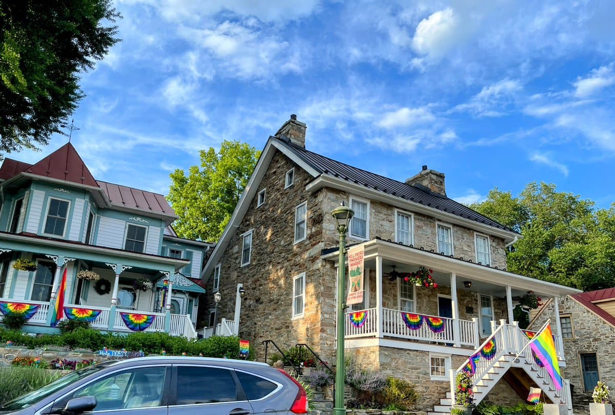 A sunny outdoor photograph of two historic buildings, one stone and one painted green, decorated with rainbow flags and banners on their porches and railings. A silver car is parked in the foreground under a blue sky with scattered clouds.