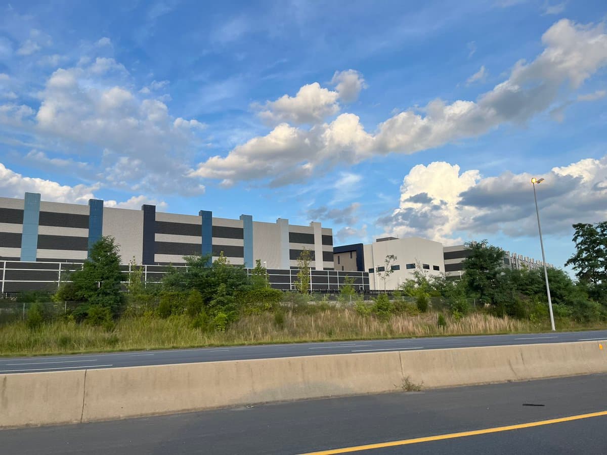 A photograph of a large, modern industrial or commercial building with a striped facade, viewed from across a highway on a sunny day with a blue, partly cloudy sky.
