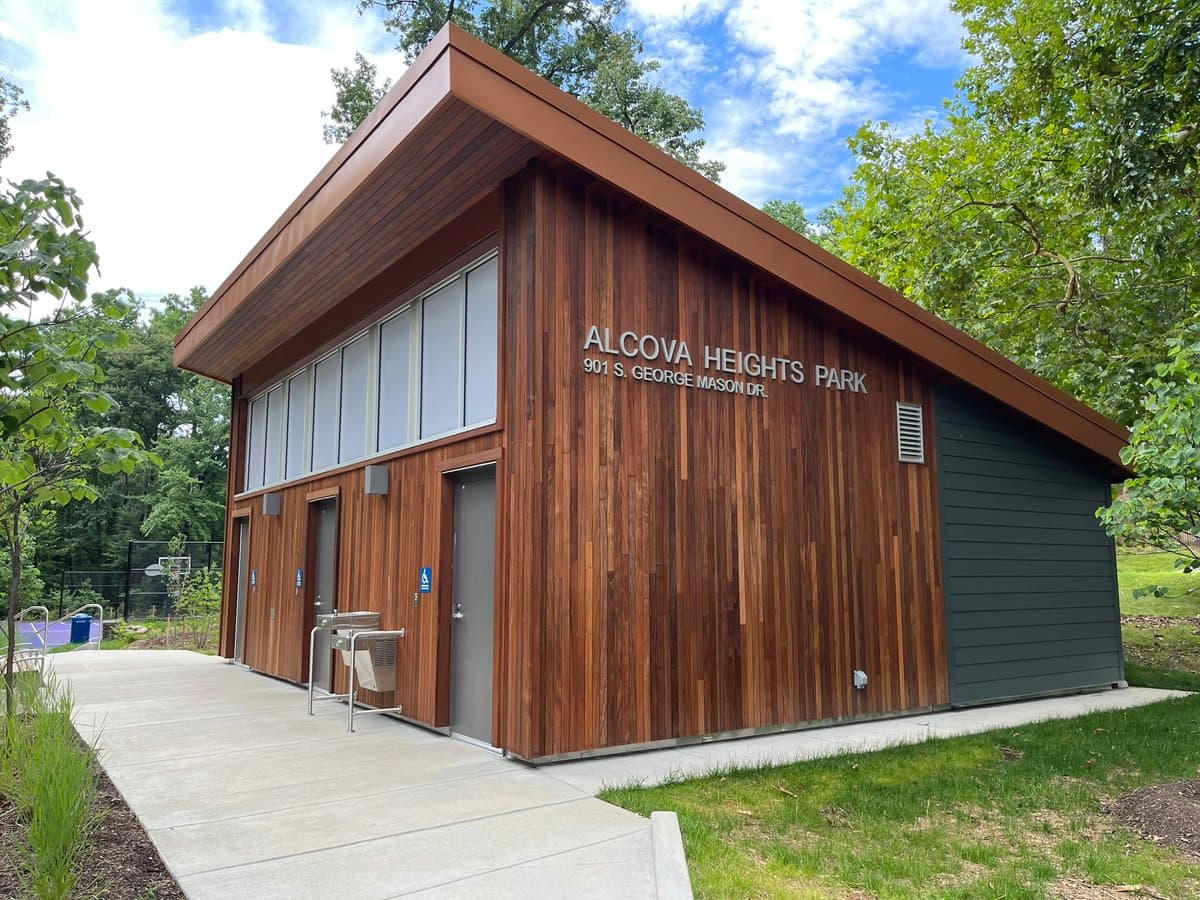 A modern, wood-paneled public restroom building with a sloped roof, situated in a park setting with trees and a concrete walkway. The structure features multiple doors, including an accessible entrance, and a sign on the side wall.