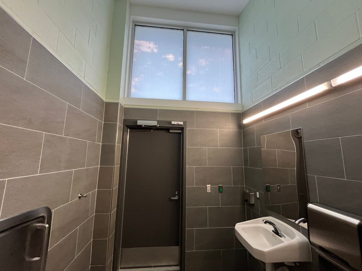 A photograph of a modern public restroom interior featuring dark grey tiled walls, a high window, a dark door, a wall-mounted sink, and a mirror.