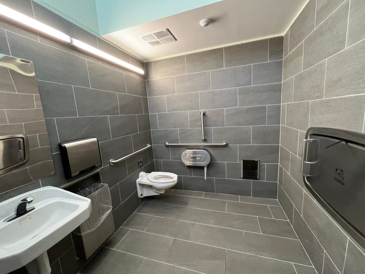 A photograph of a modern, accessible bathroom featuring grey tiled walls and floor, a wall-mounted toilet, a sink, and multiple safety grab bars.