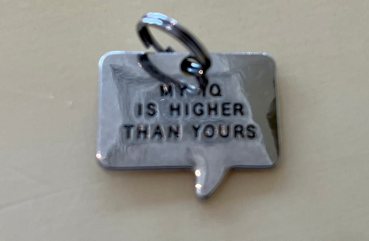 A close-up photo of a silver, speech-bubble-shaped metal tag with a split ring, intended as a pet collar charm. The tag is engraved with the text "MY IQ IS HIGHER THAN YOURS" in capital letters.