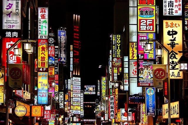 A nighttime photograph of a densely packed urban street, likely in Japan, illuminated by numerous colorful neon signs and billboards in Japanese. The scene features a vertical composition with a dark sky in the center, surrounded by glowing advertisements for various businesses.