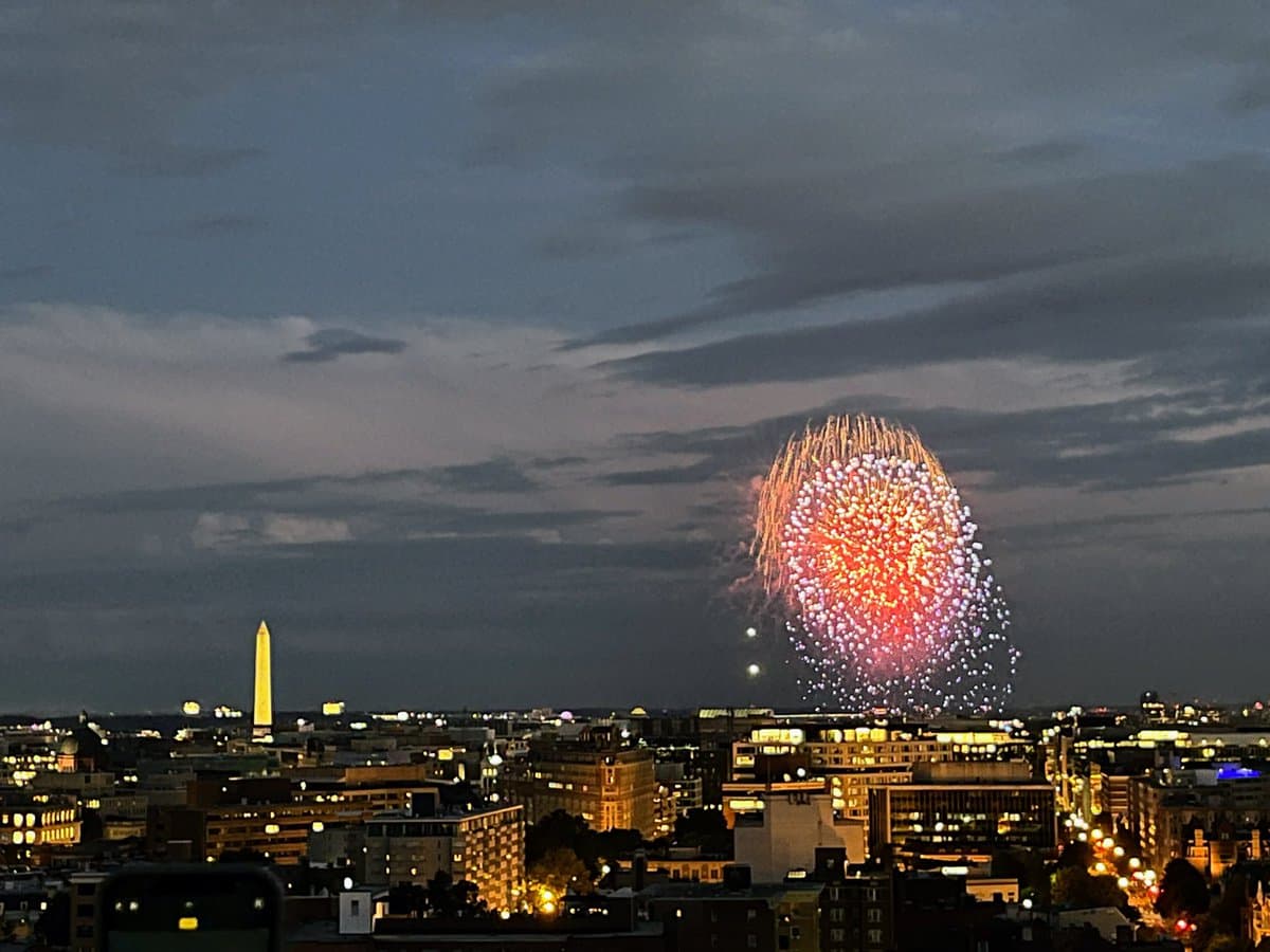 A nighttime cityscape featuring a large, colorful firework exploding in the sky above a dense urban skyline. The Washington Monument is visible on the left side of the horizon, illuminated against the dark, cloudy sky.